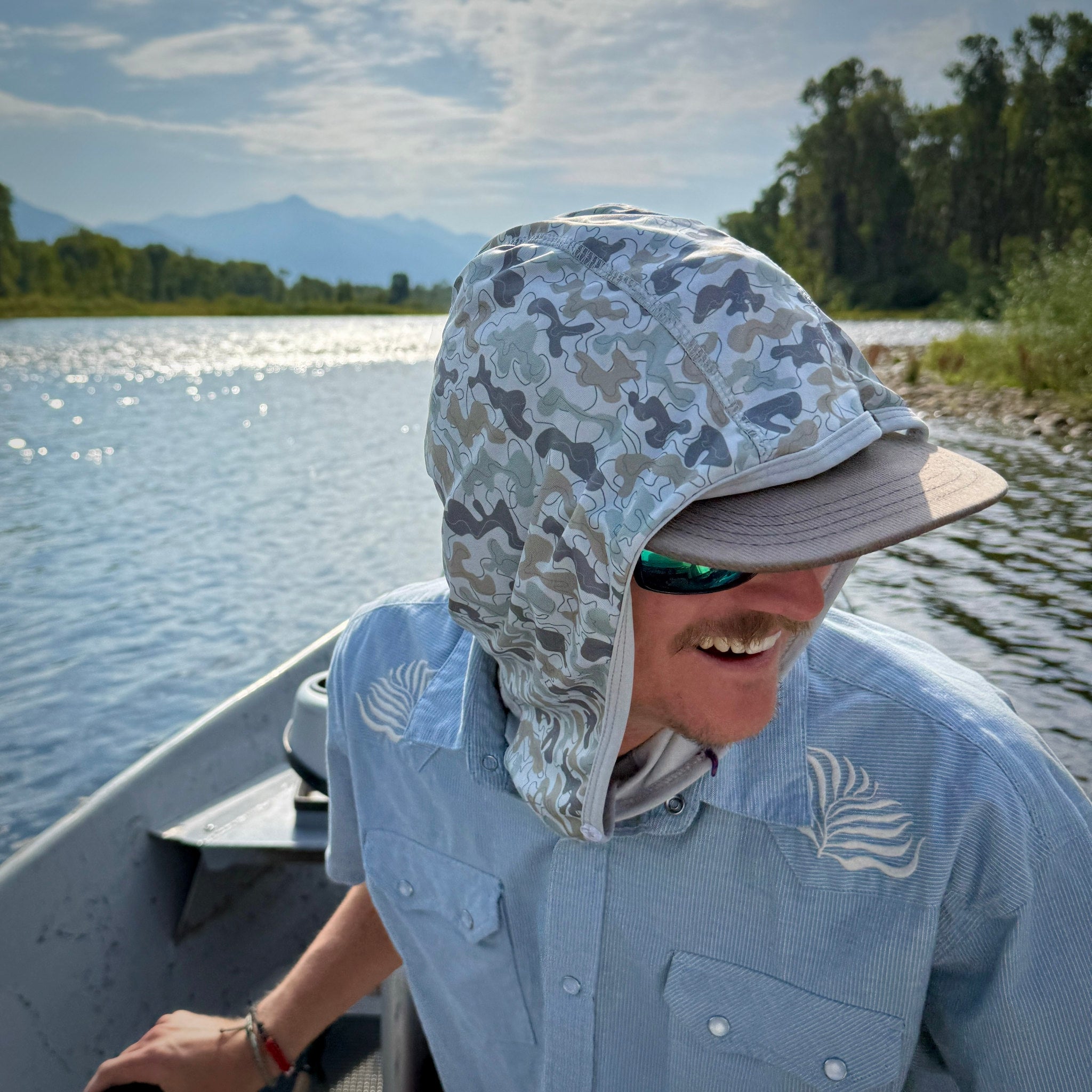 Young man on a boat wearing a camouflage sun hood and sunglasses with a scenic background.