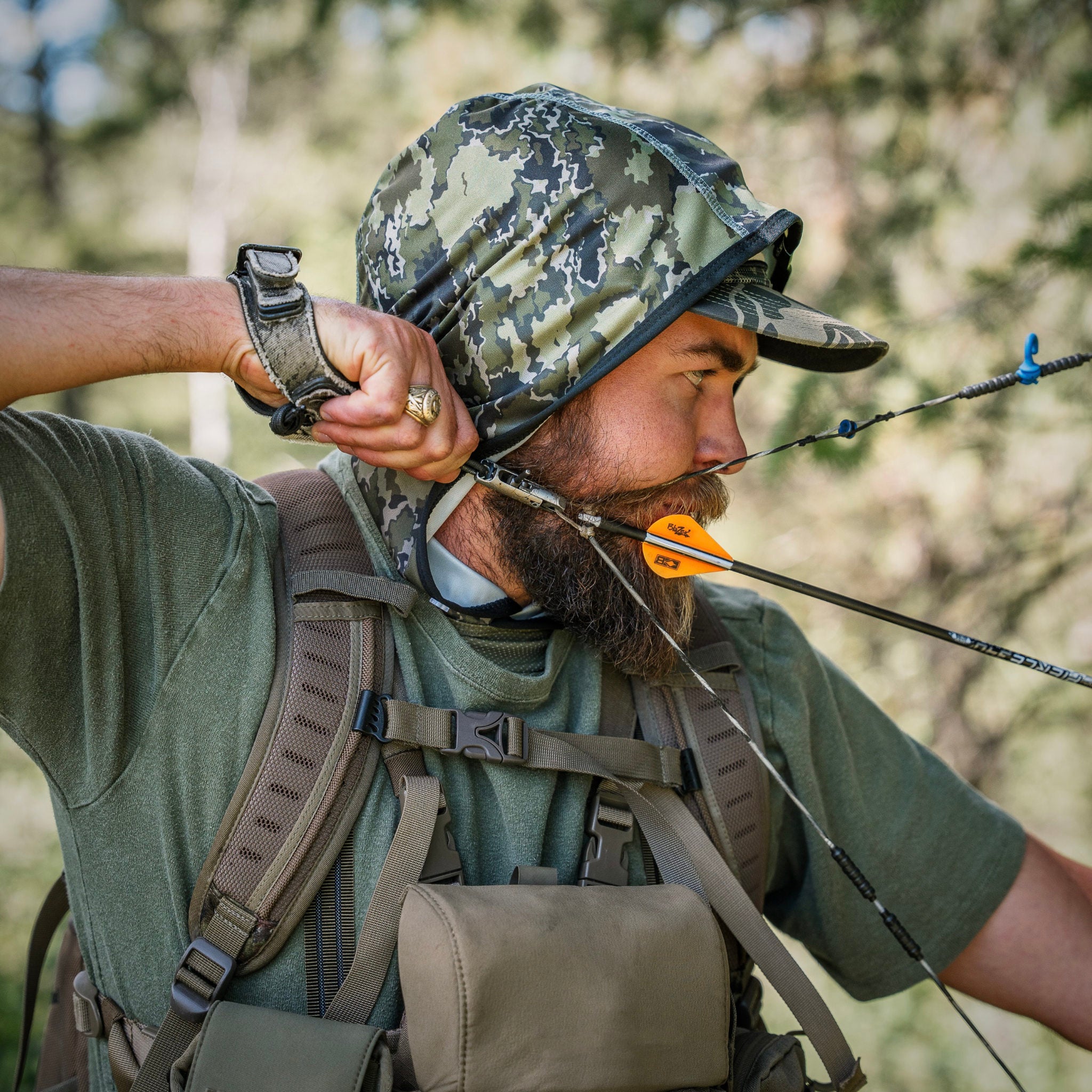 A man in an olive green shirt and camouflage hunting Kinich Sun Hood draws a bow while wearing a backpack in an outdoor forest setting.